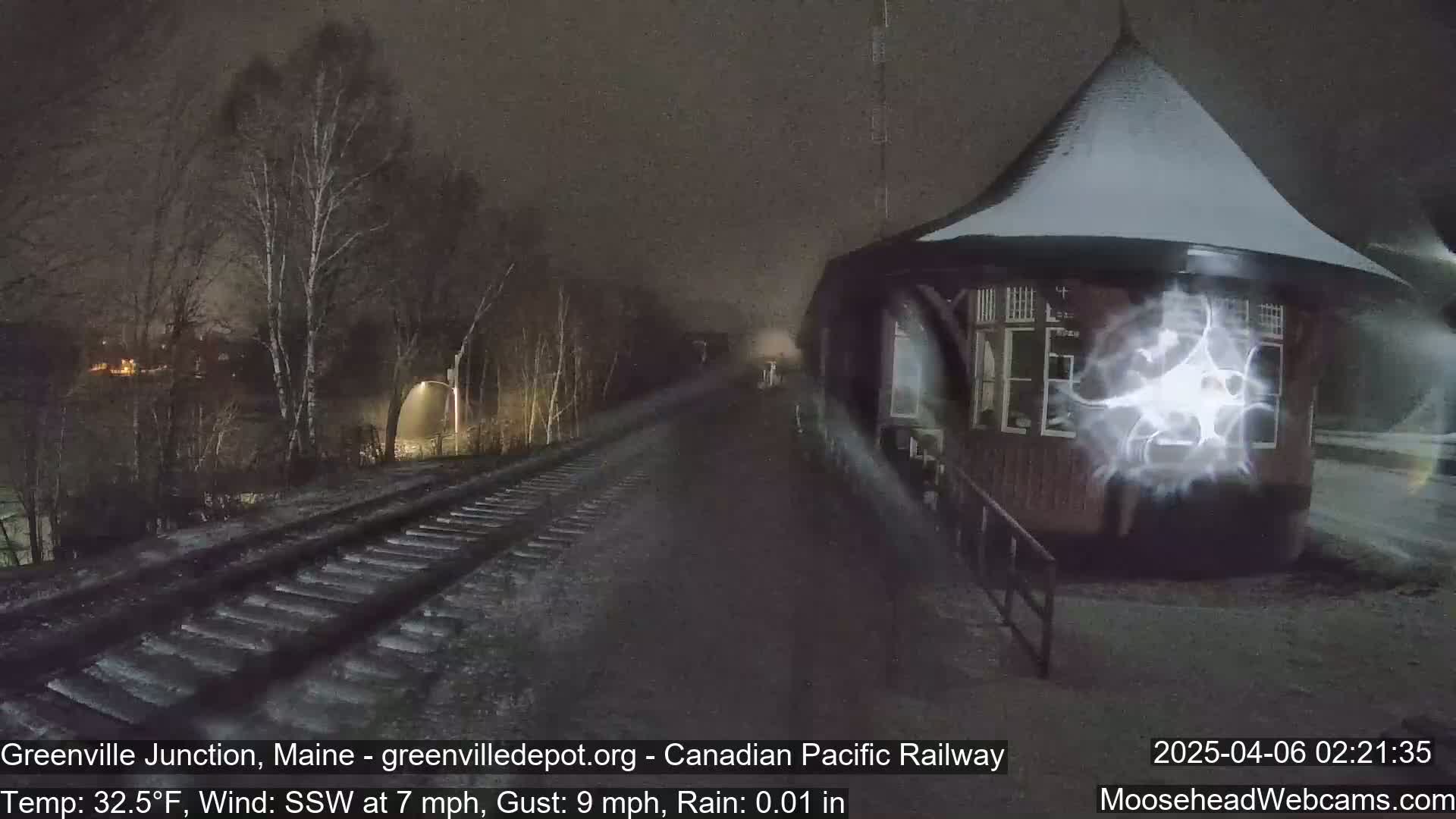 A snow-covered, dark-brown gazebo sits beside a railroad track in the rain at night, with bare trees and dimly lit buildings in the background.