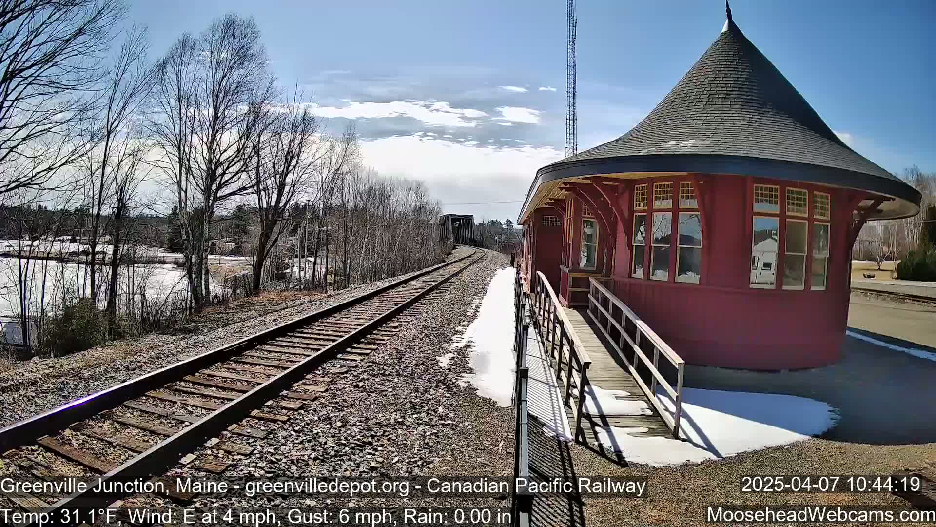 A red, octagonal train station with a conical roof stands beside railroad tracks on a partly sunny day with patches of snow and ice on the ground.