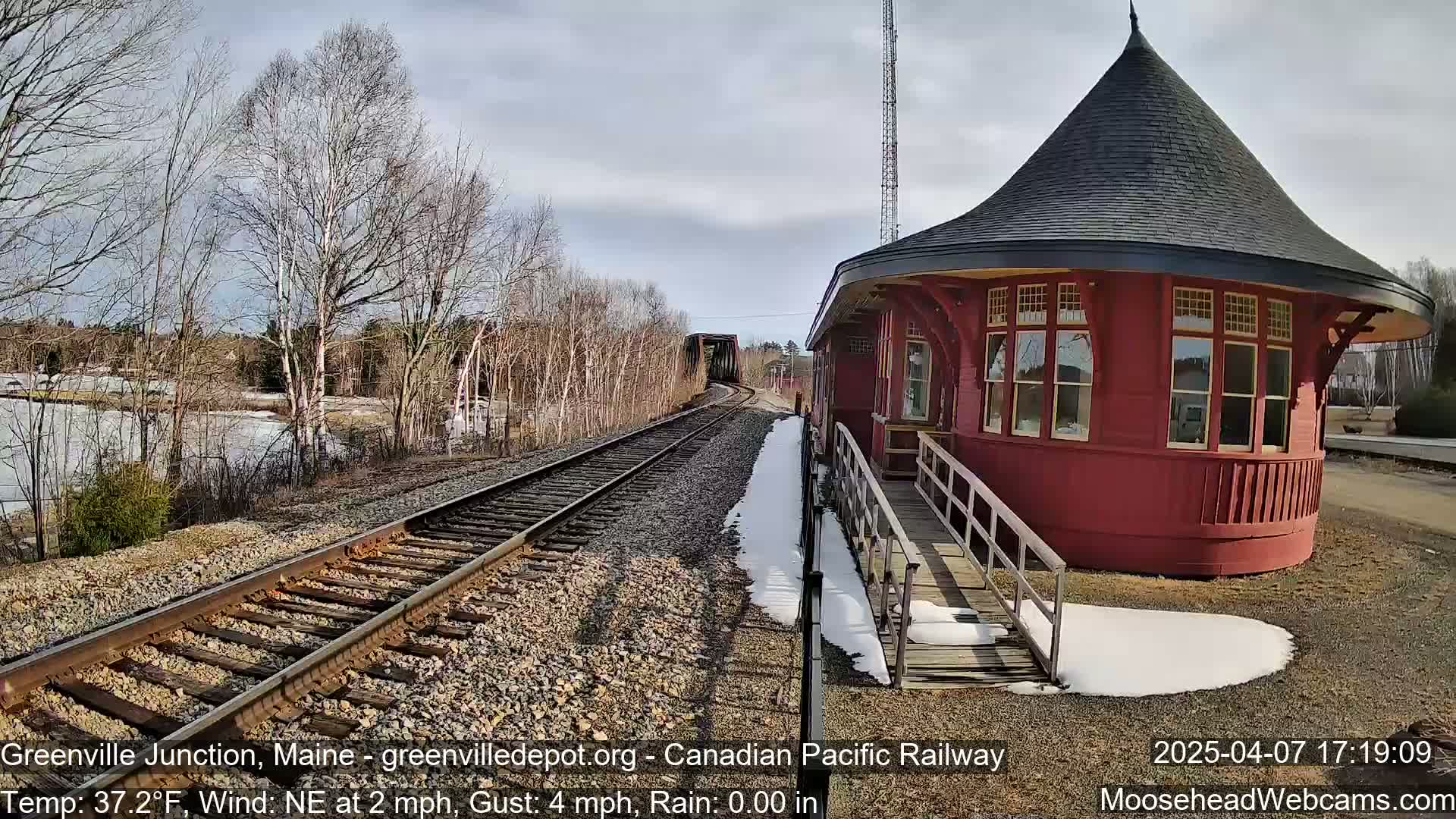 A red, round train depot with a conical roof stands next to train tracks on a partly sunny day with patchy snow on the ground and some ice on a nearby body of water.