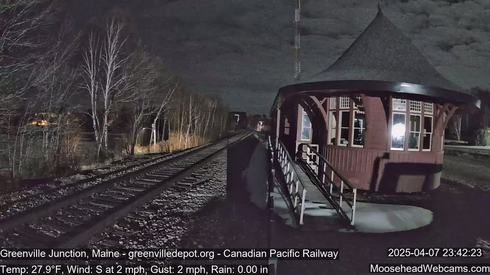 A dark reddish-brown train depot with a conical roof stands beside train tracks at night under a cloudy sky in light rain.
