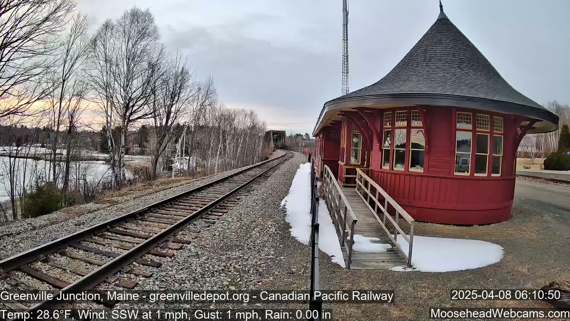 A red, round train station with a conical roof sits beside railroad tracks, partially covered in snow, under a mostly cloudy sky.
