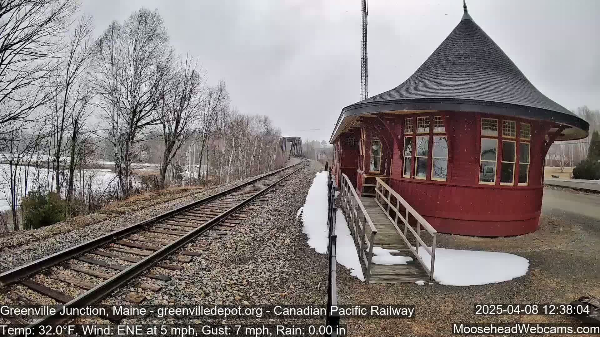 A red, round train depot with a dark roof sits next to a railway track, partially covered in snow, under a gray, snowy sky.