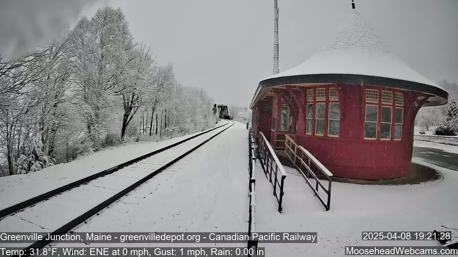 Snow falls on a snow-covered railway scene with a red, round train station and trees.