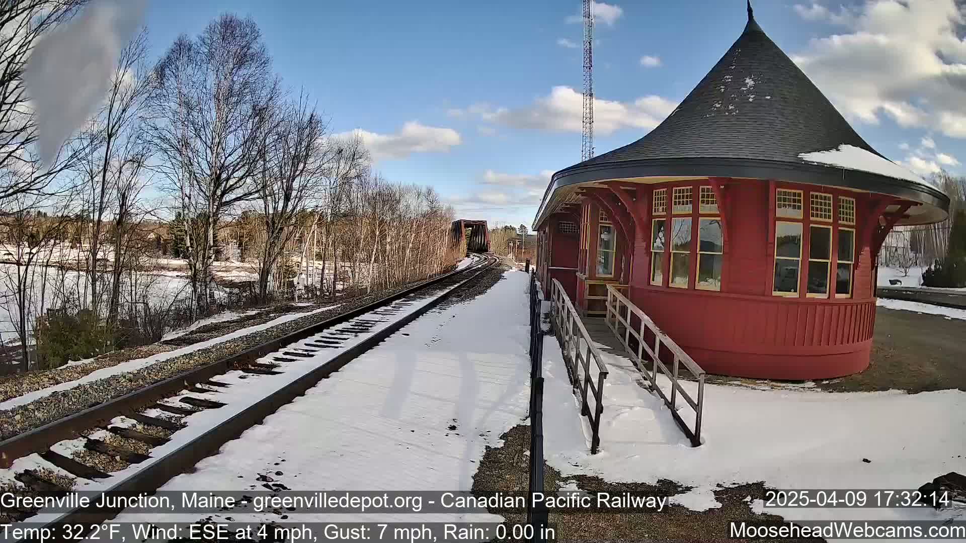A red, round train depot sits beside snow-covered train tracks on a partly sunny day with a light wind.