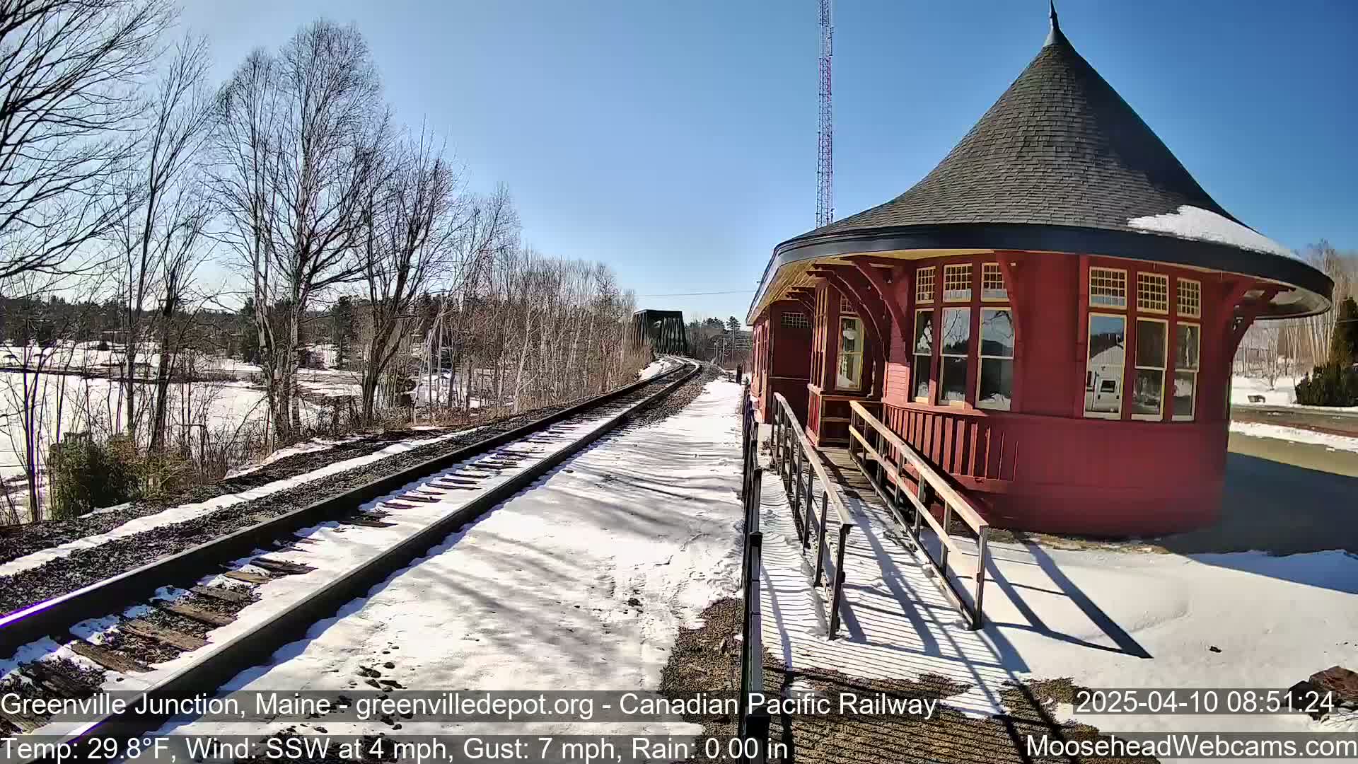 A red octagonal train station sits beside snow-covered train tracks on a sunny day with a light south-southwesterly wind.