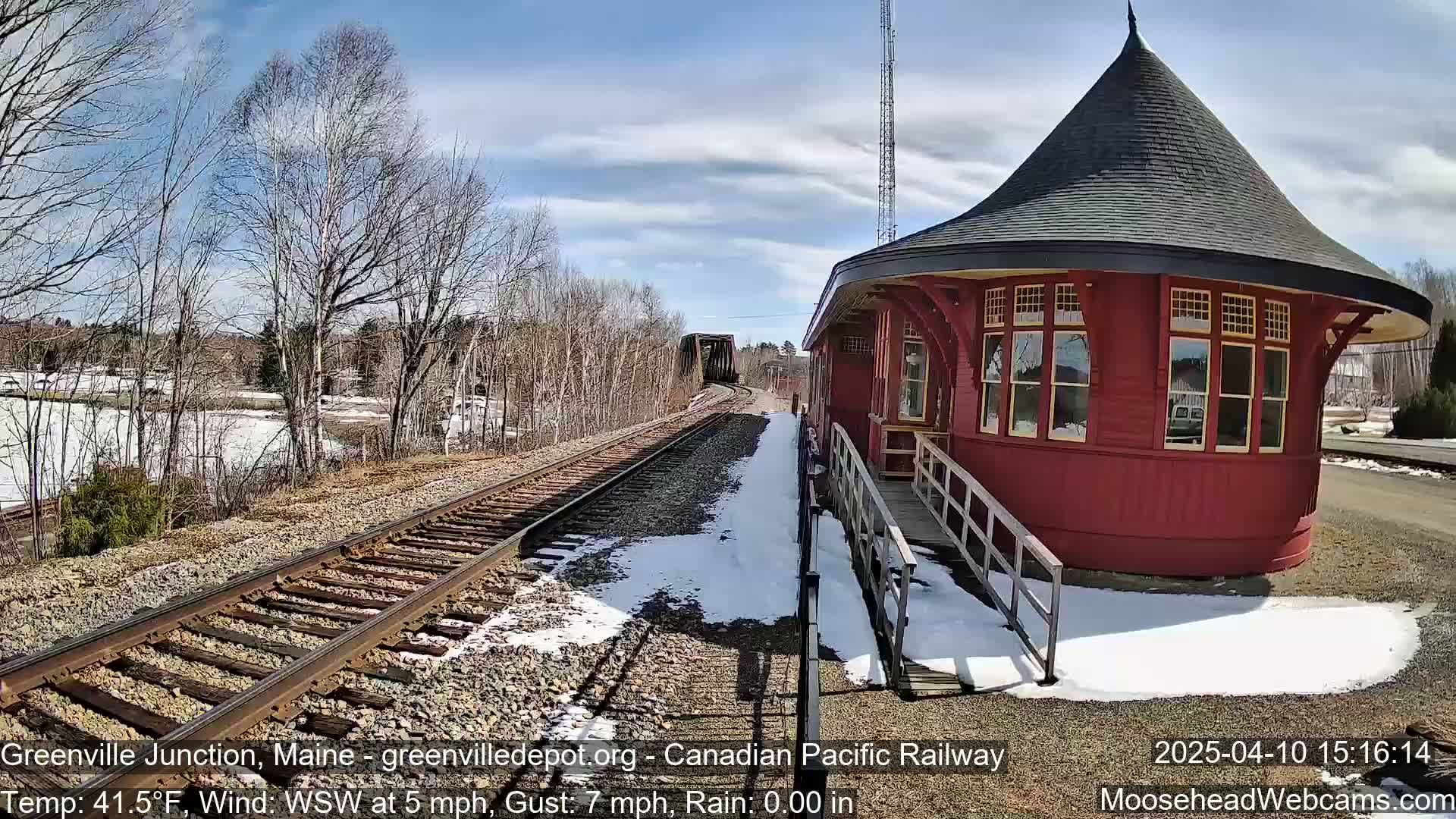 A red, round train station with a dark conical roof sits next to train tracks partially covered in snow on a partly sunny day.