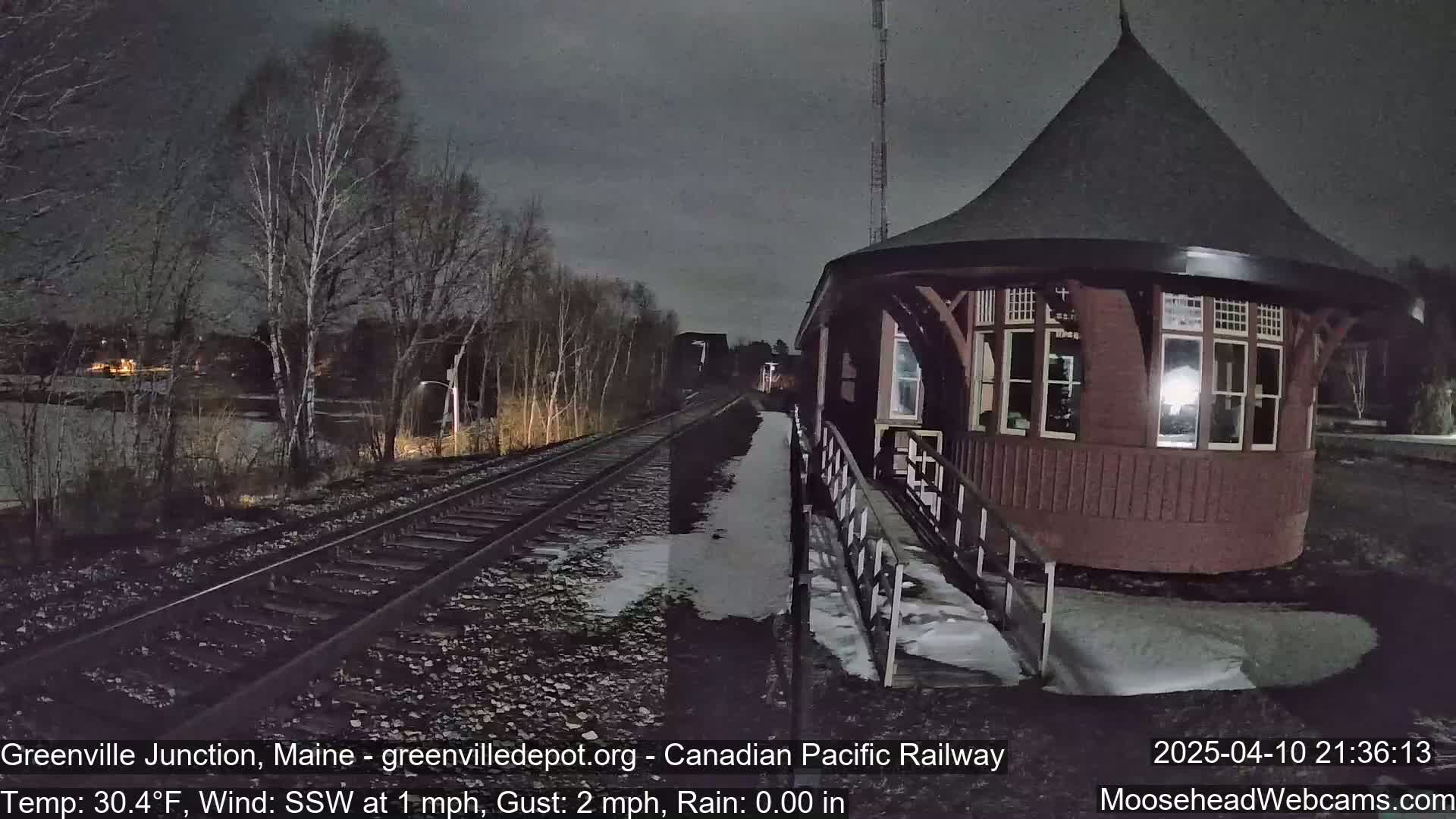 A dark, rainy night scene shows a railway track leading to a round, red train station building with patches of snow and ice on the ground.