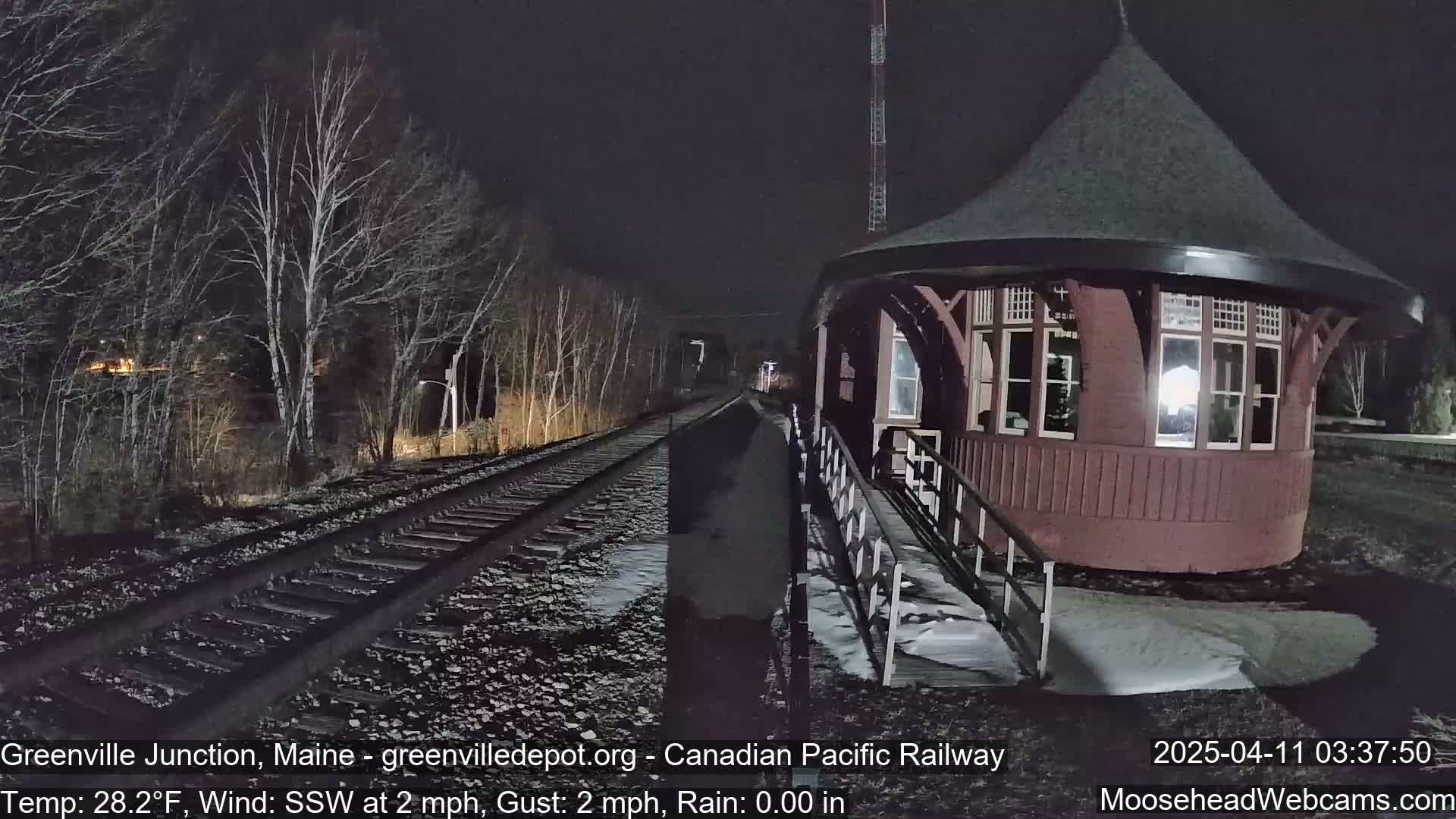 A dark nighttime scene shows a train track leading to a reddish-brown train station with a conical roof, under a light rain with a temperature of 28.2°F and a south-southwesterly wind.