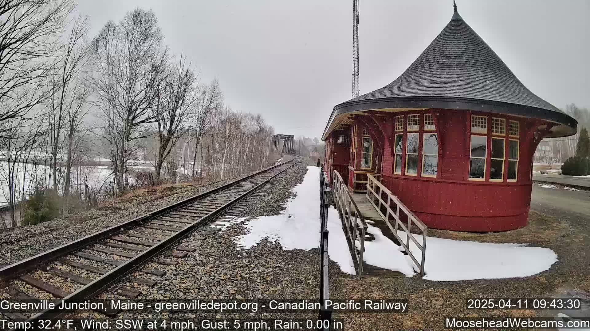 Snow falls on a red, round train depot next to a set of train tracks leading to a bridge, with bare trees and a partially snow-covered landscape in the background.