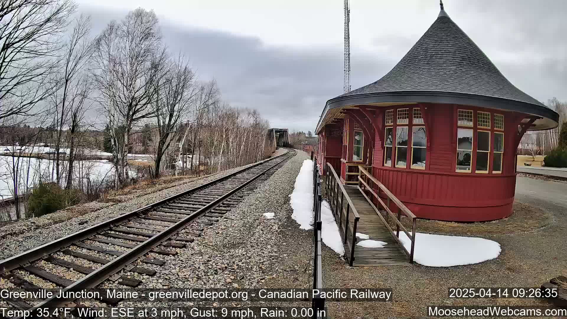 A red octagonal train depot with a conical roof sits beside train tracks on a partly cloudy day with patches of snow on the ground.