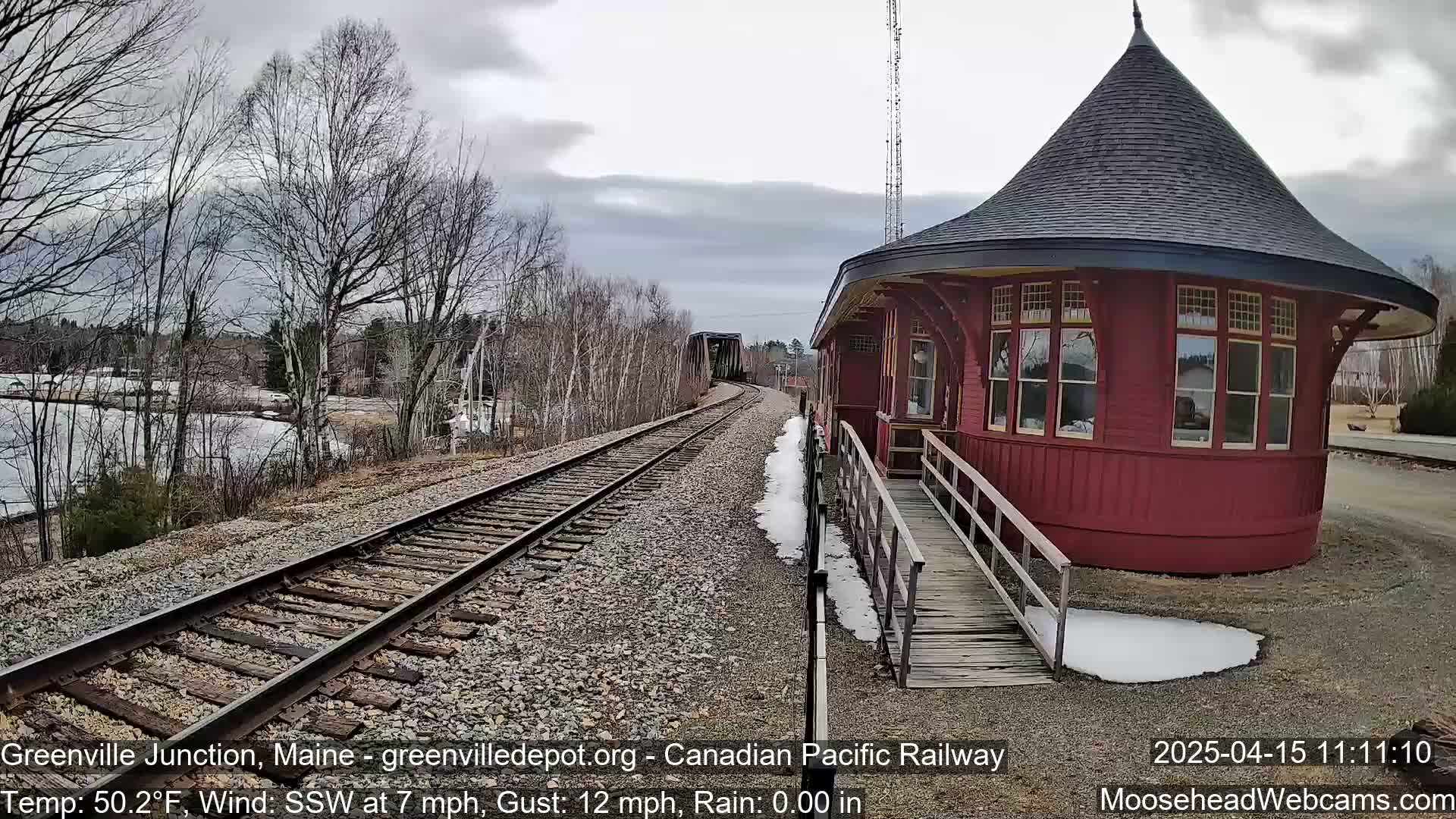 A red, round train station with a conical roof stands next to railway tracks leading to a bridge, under a cloudy sky with light rain and patches of snow on the ground.
