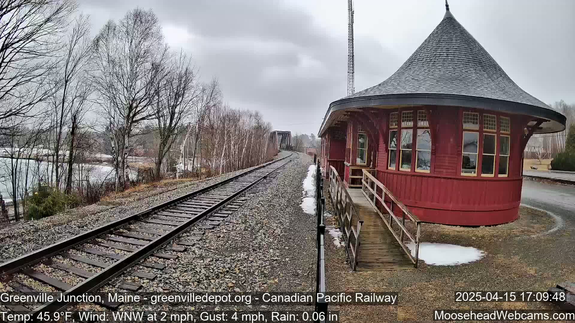 A red octagonal train depot sits beside railway tracks extending to a bridge under a gray, rainy sky.