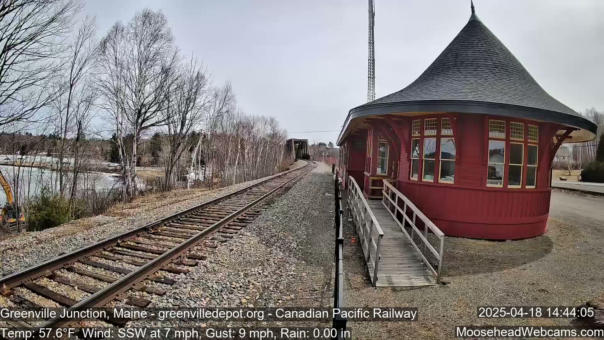 A red, round train depot with a conical roof stands beside railroad tracks that stretch towards a bridge, under a partly cloudy sky.