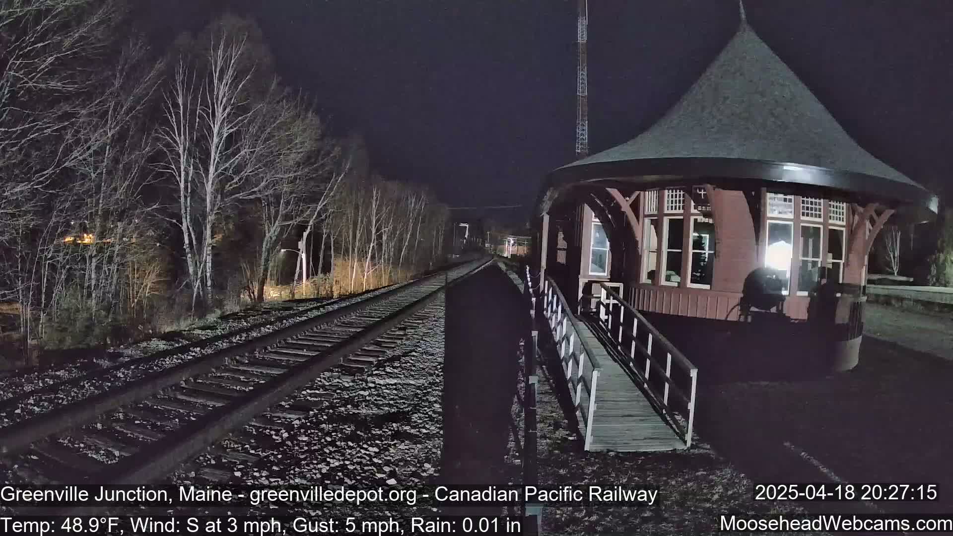 A dimly lit train depot with a ramp stands next to railroad tracks under a dark, rainy night.