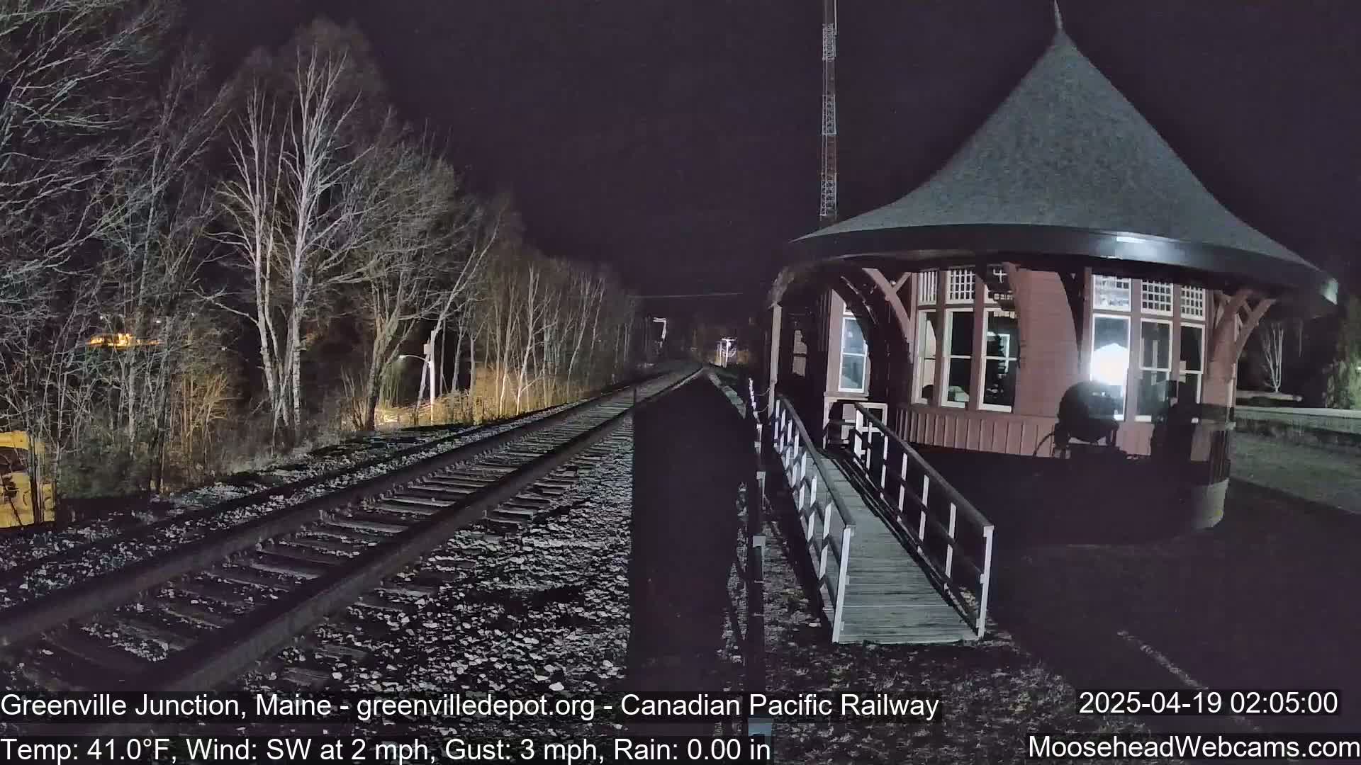 A nighttime view of train tracks leading to a round, brown railway station building beside bare trees under a dark sky.