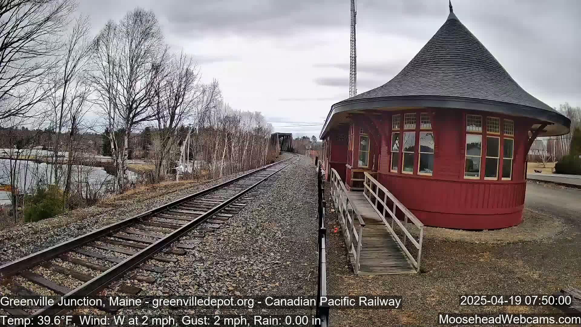 A red, round train depot with a conical roof stands beside railroad tracks leading to a bridge under a cloudy sky.