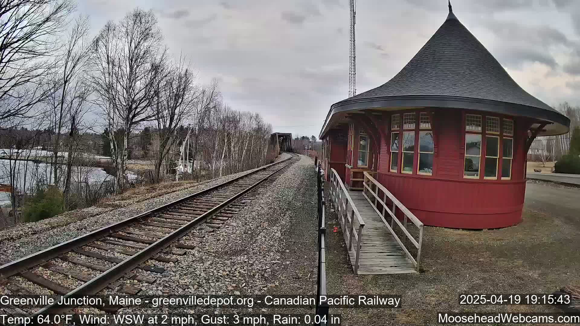 A red, round train depot with a conical roof stands beside railroad tracks leading to a metal bridge, under a cloudy sky with light rain.