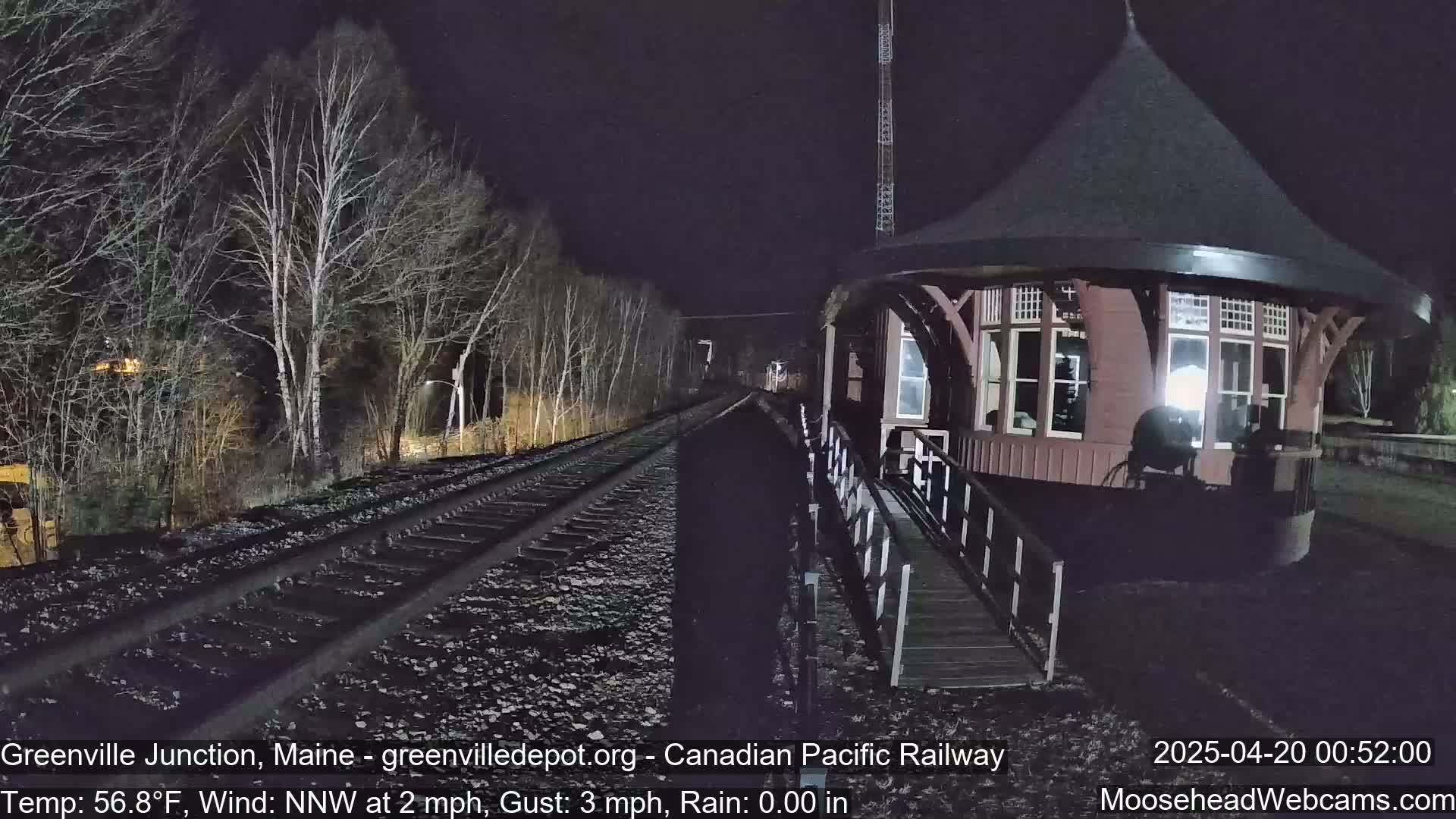A nighttime view of a train station with a ramp leading to it, situated beside railroad tracks that disappear into a dark, sparsely wooded area under a rainy sky.