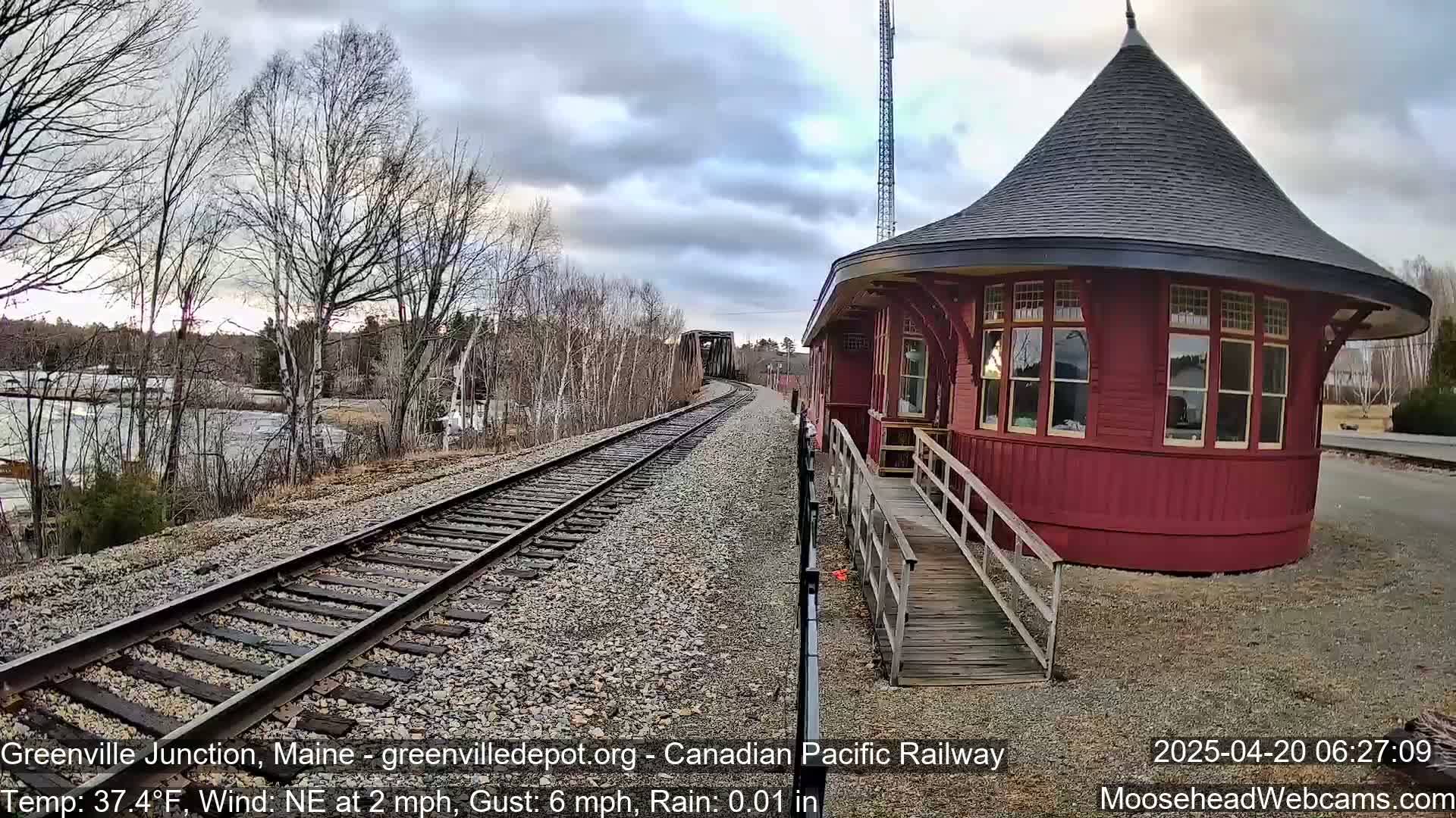 A red, round train depot with a conical roof stands beside railroad tracks on a partly cloudy day with light rain and a northeast wind.