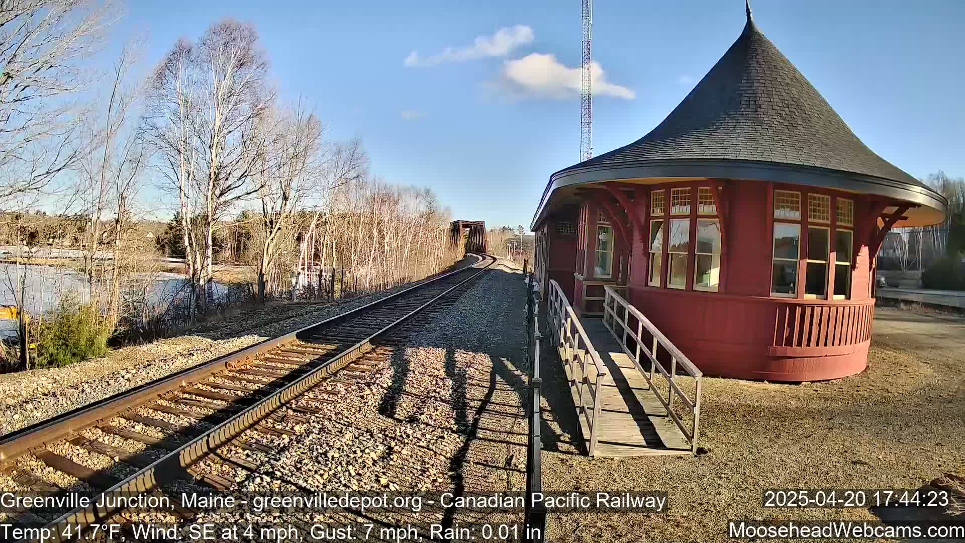 A red, octagonal train station with a conical roof sits next to railroad tracks that extend to a bridge under a mostly sunny sky with a light south-easterly wind and a trace of rain.
