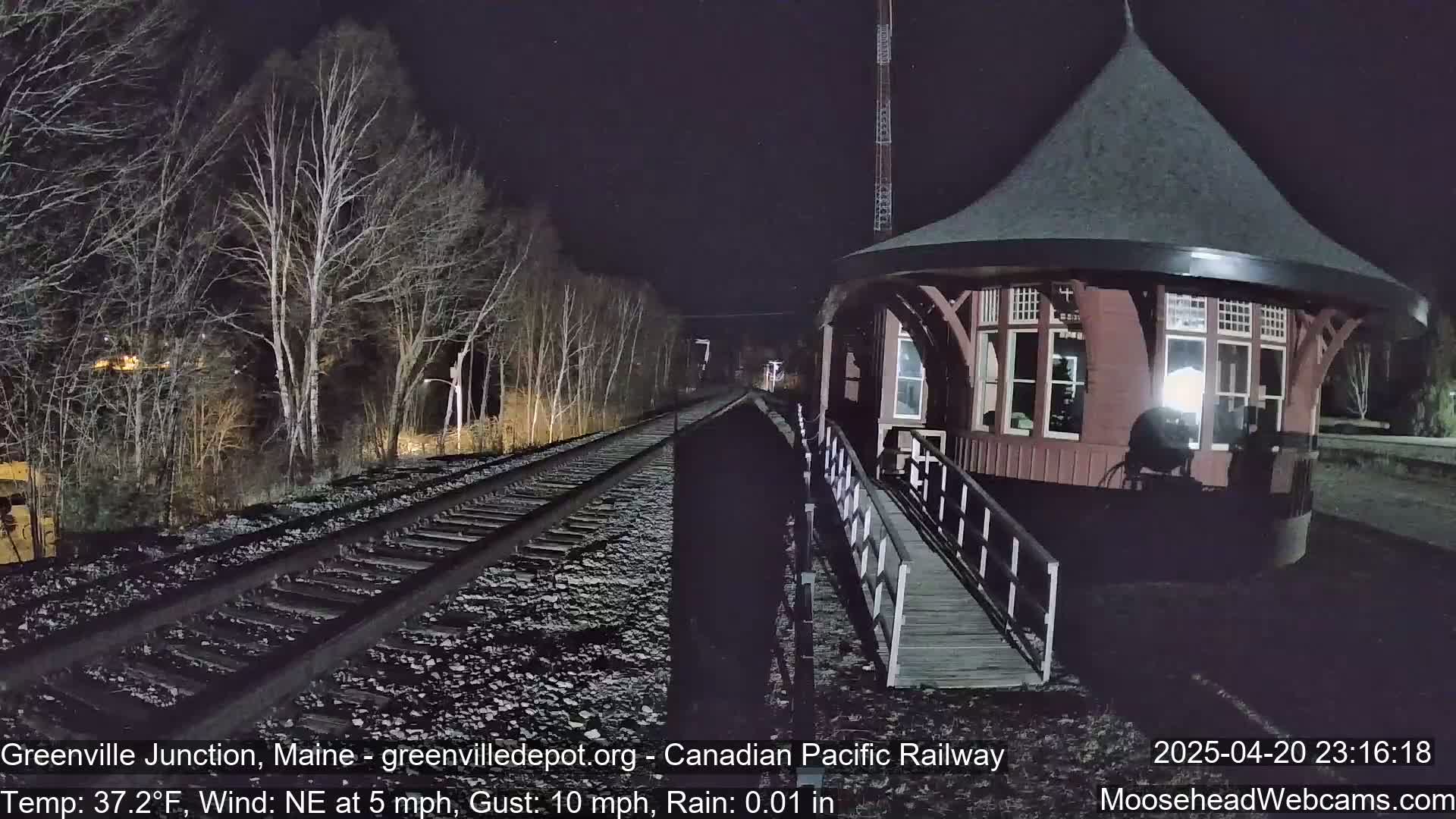 A nighttime view of a railway track leading to a round, red train station under a lightly rainy sky.