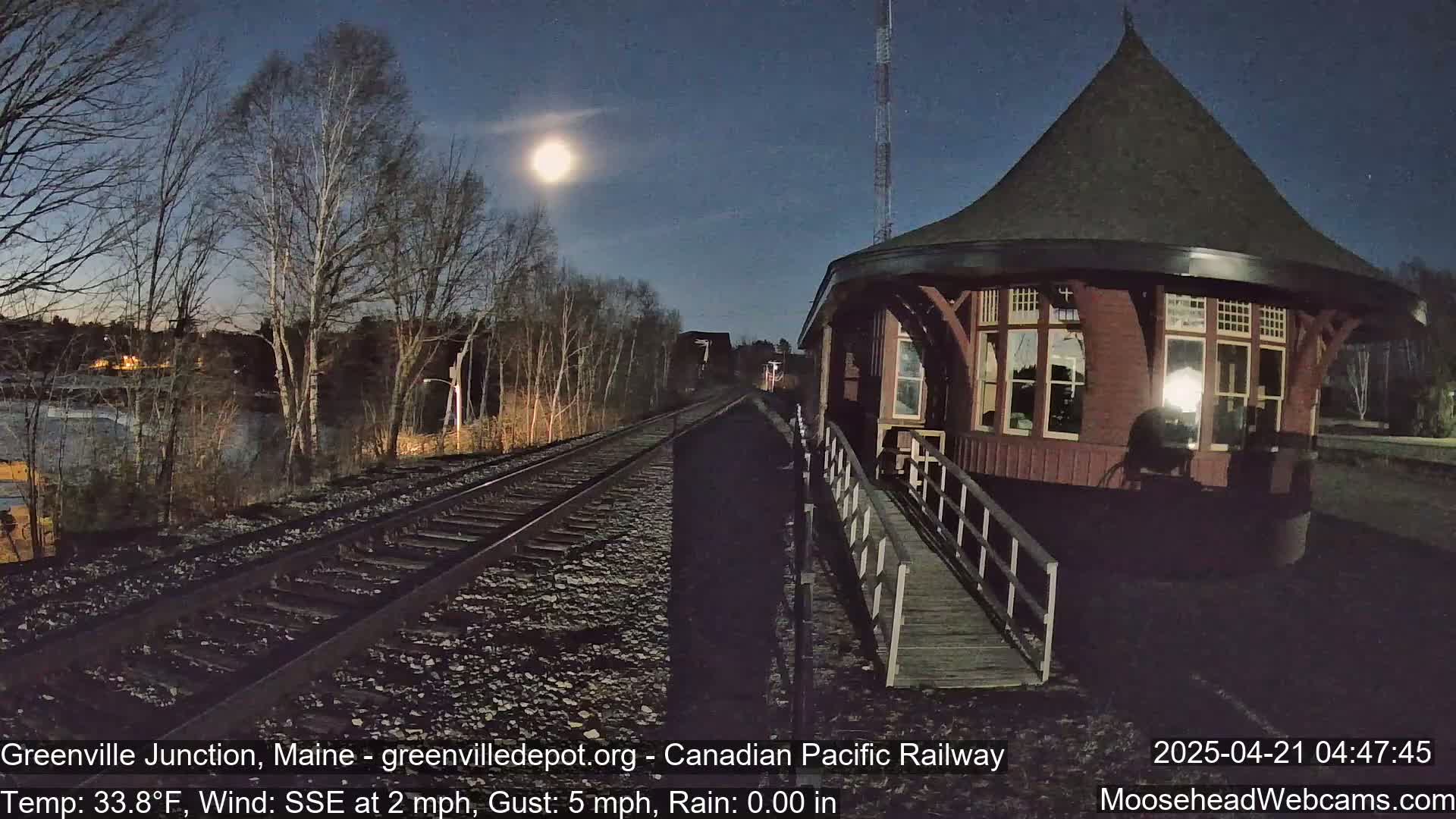 Under a bright moonlit sky, a red train depot stands beside railroad tracks, with bare trees and a calm body of water visible in the background.