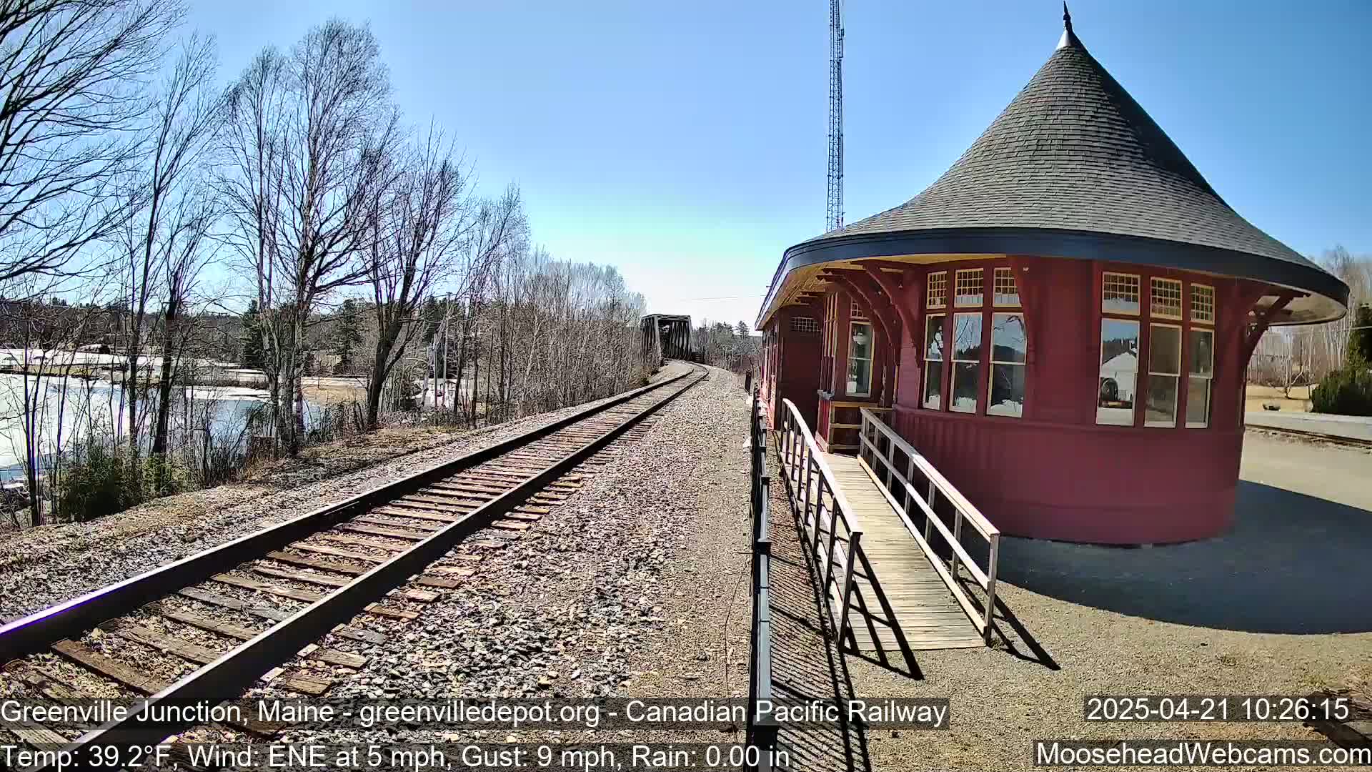 A red, round train station with a conical roof sits beside railroad tracks leading to a bridge under a sunny sky with a light wind.