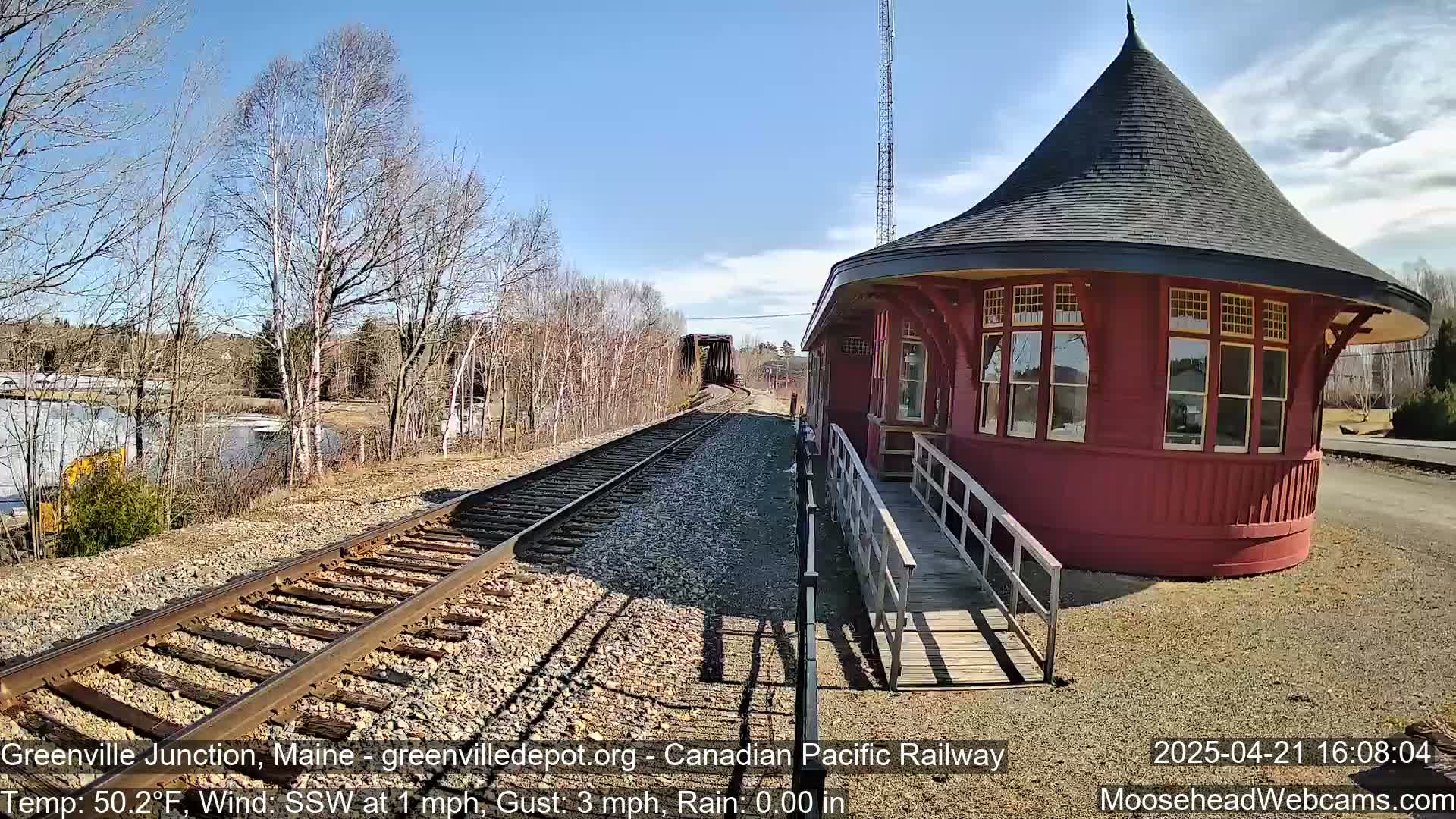 A red, round train depot with a conical roof sits beside railroad tracks on a sunny day, with a body of water and bare trees visible in the background.