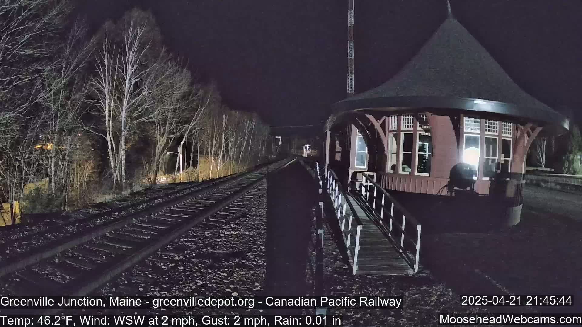 A dark, rainy night scene shows a red, octagonal train depot next to railroad tracks that wind through bare, leafless trees.