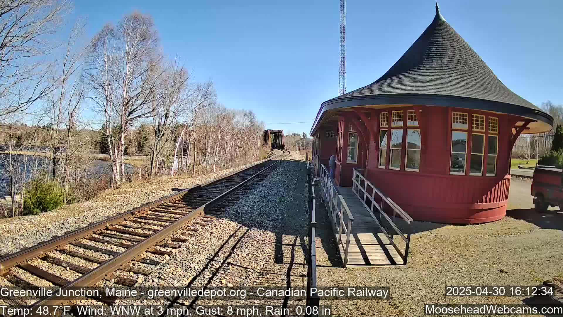 A red, round train depot with a conical roof sits beside railroad tracks that extend to a bridge over a body of water under a sunny sky with light rain.