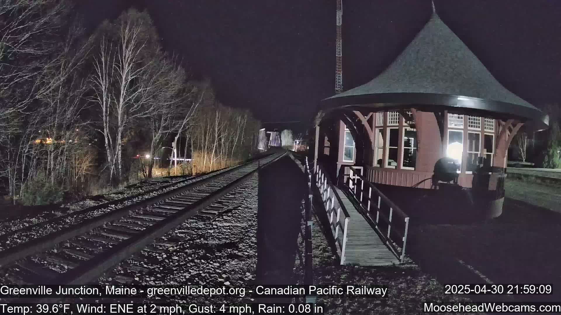 At night, under rainy conditions, a red train depot is next to railroad tracks that extend into a dark, wooded area.