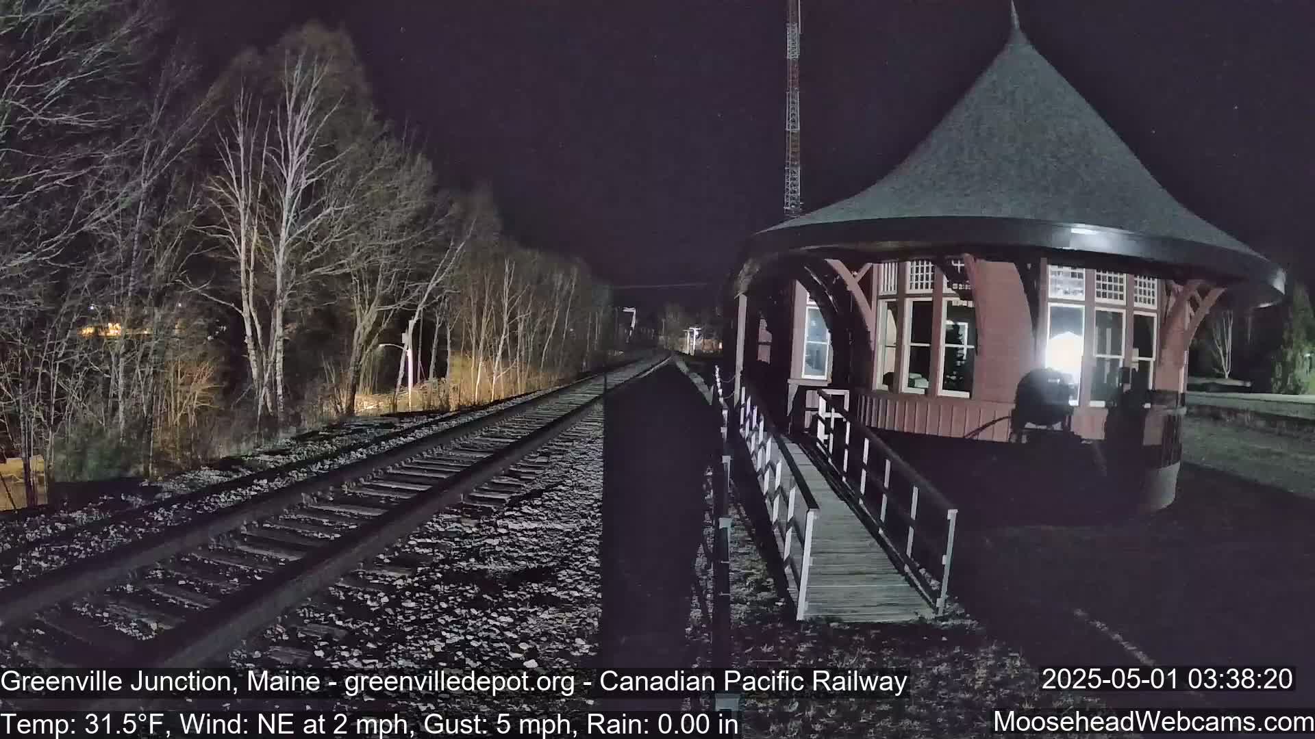 A dark, nighttime scene shows a train track leading to a round, reddish-brown railway station building next to bare trees under a clear sky with a northeast wind.