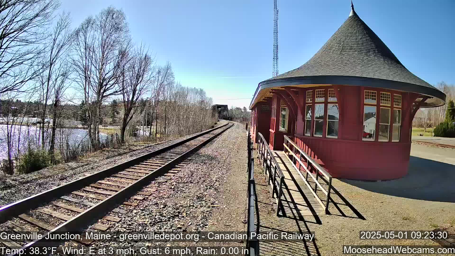 A red, round train station with a conical roof sits beside railroad tracks on a sunny day with a light east wind.