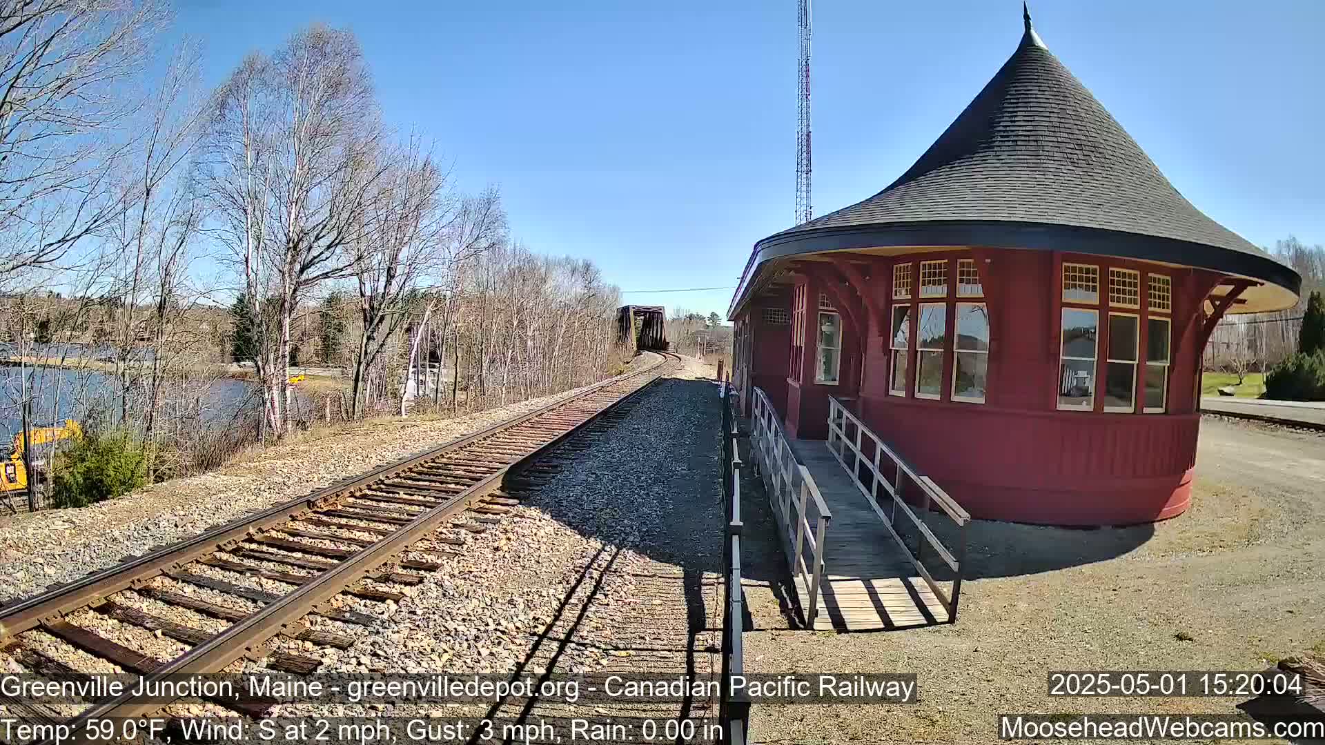 A red, round train depot with a conical roof sits next to railroad tracks leading to a bridge over a body of water under a sunny sky.