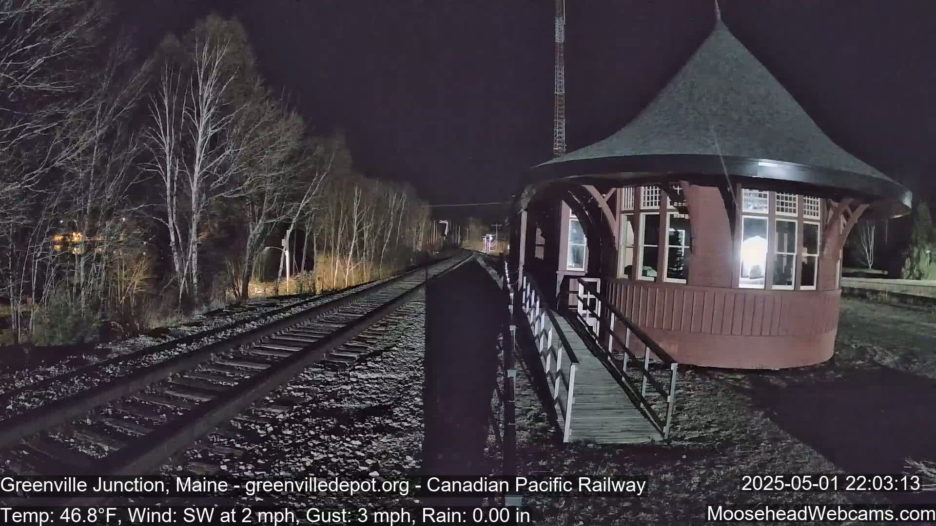 A dark, nighttime view shows a round, brown train depot next to train tracks that run through a sparsely wooded area, with a light southwest wind and a temperature of 46.8°F.