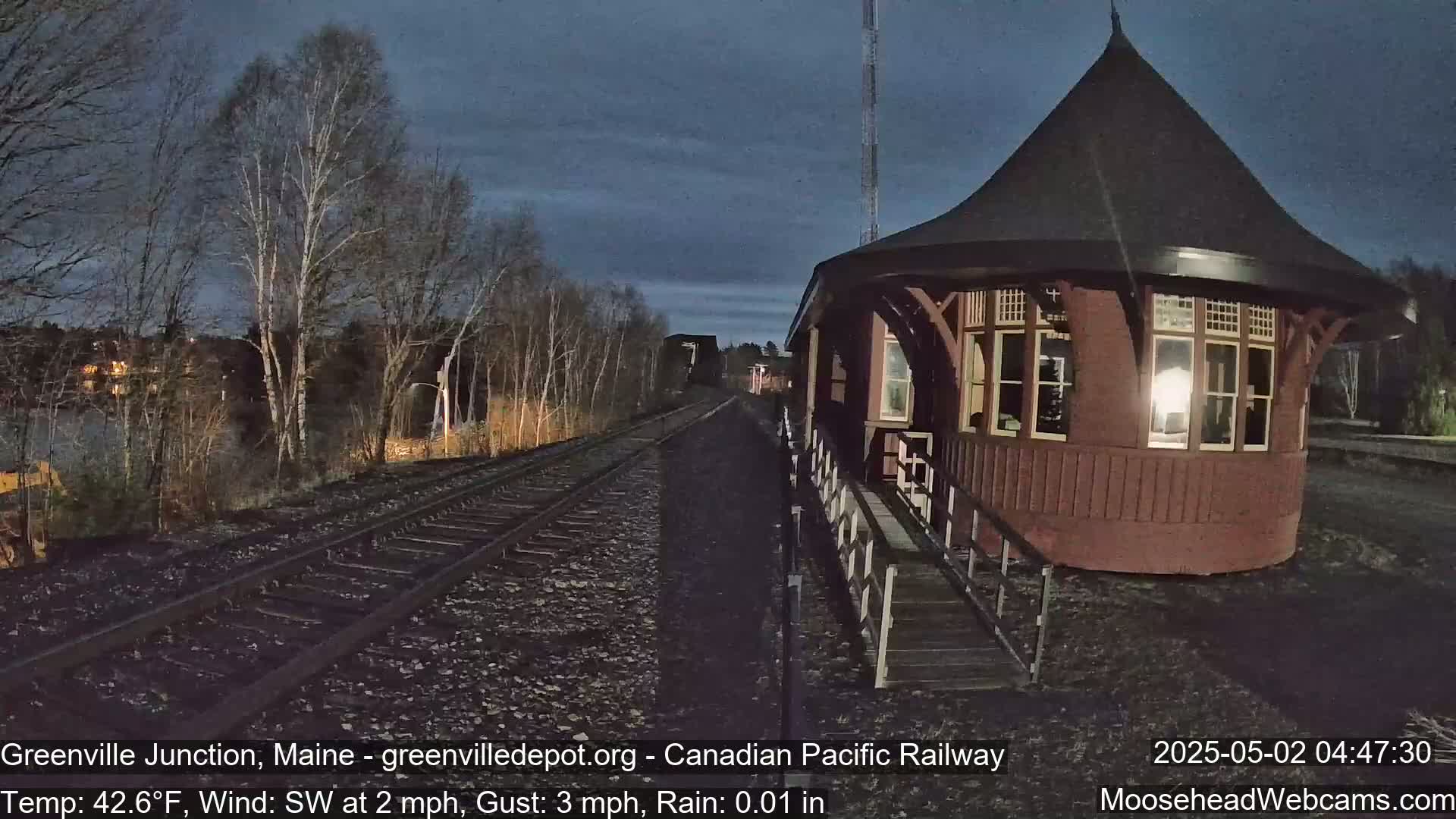 A red train station with a conical roof stands beside railroad tracks under a dark, rainy sky at night, with bare trees in the background.