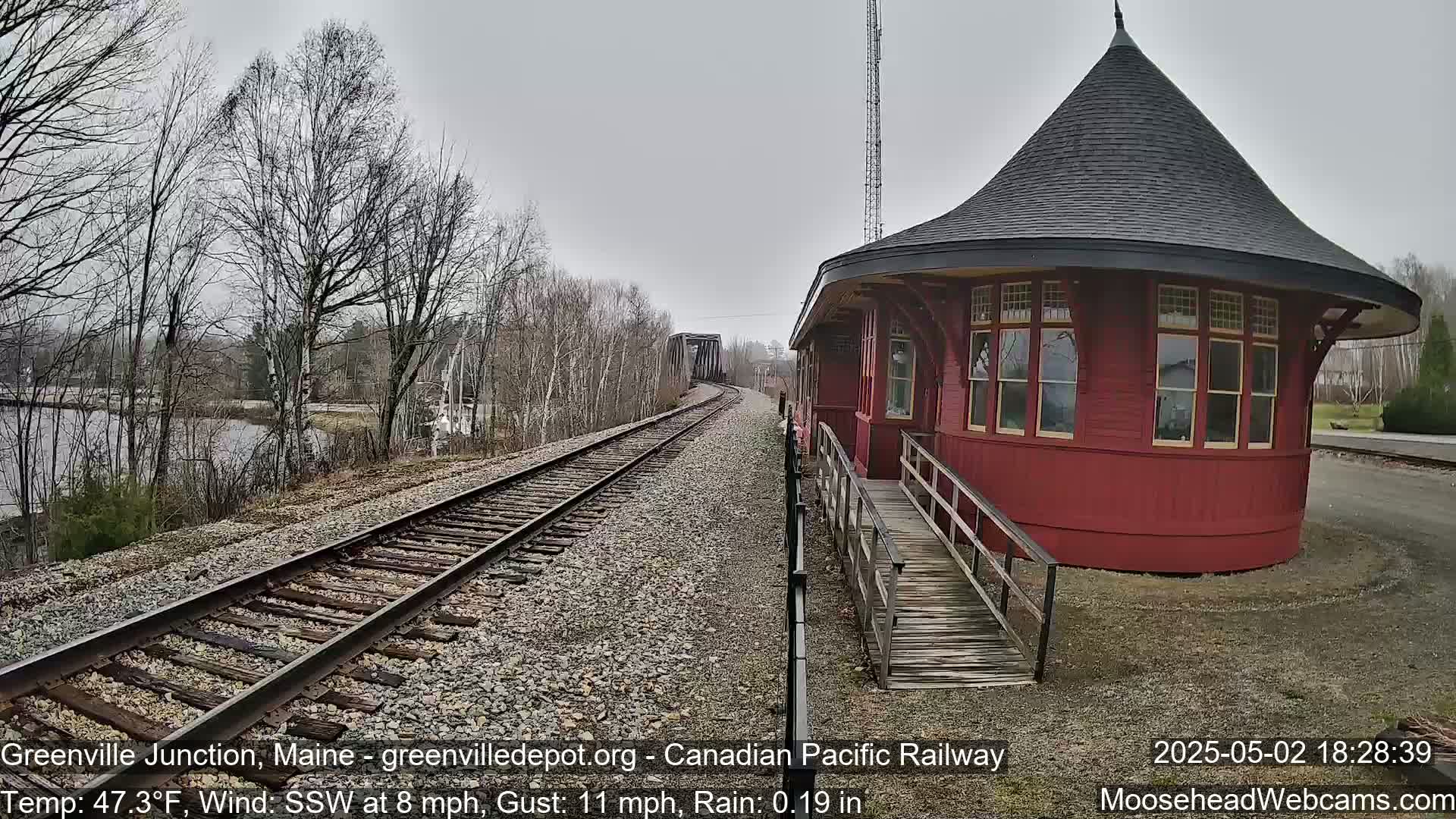 A red, round train depot with a ramp sits beside railway tracks leading to a bridge, under a gray, rainy sky.