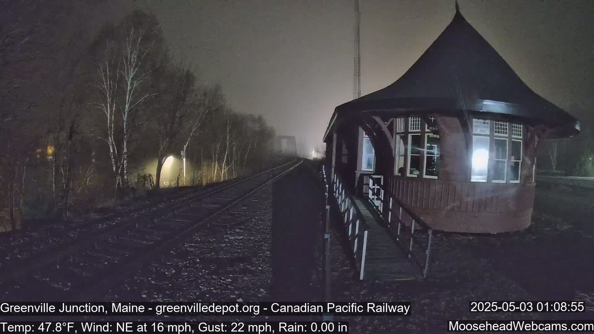 A dark, foggy nighttime scene shows a train depot next to railway tracks that recede into the distance under a hazy sky.
