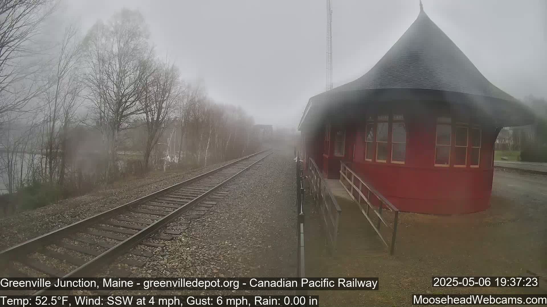 A red, round train station stands beside railroad tracks on a foggy day with a light wind.