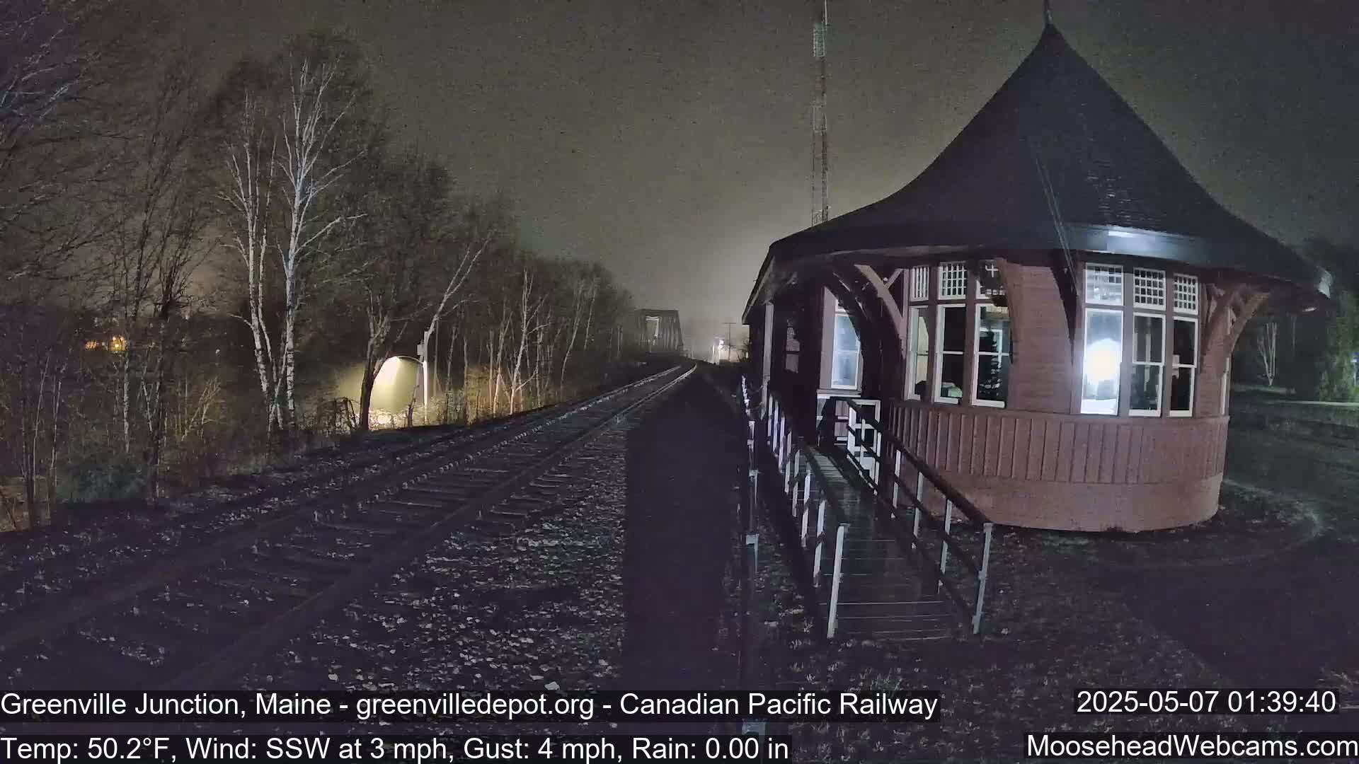 A dark, rainy night scene shows a railway track leading to a distant bridge, next to a reddish-brown octagonal railway station building under dim lighting.