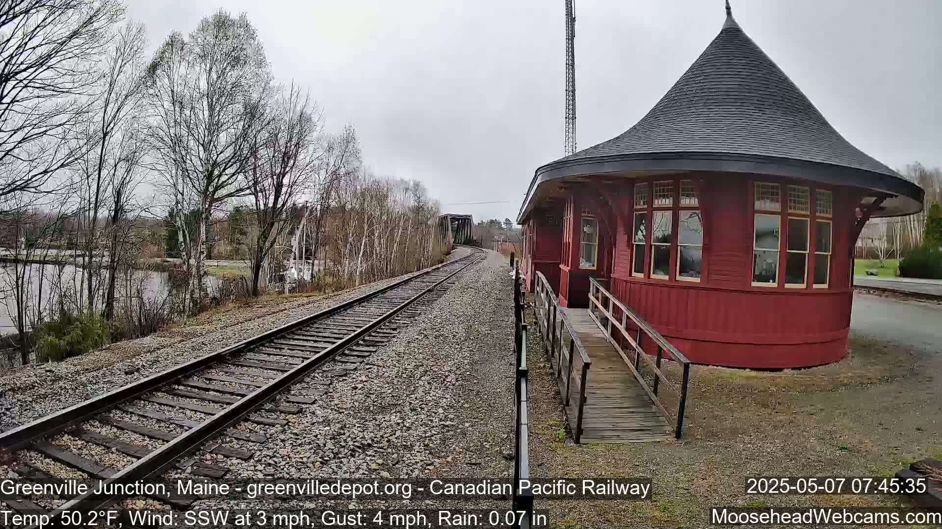 A red, round train depot with a conical roof sits beside train tracks that lead to a bridge over a calm body of water under a gray, rainy sky.