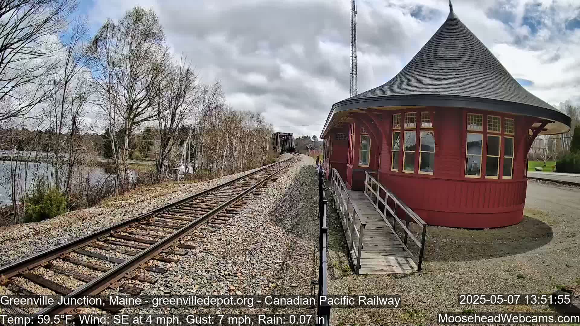 Train tracks curve toward a red, round train depot under a partly cloudy sky with light rain.