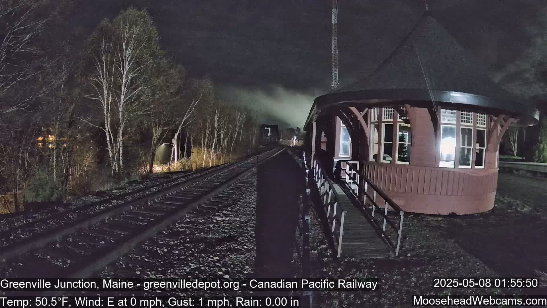 A dark, nighttime view of a train track leading to a red octagonal building next to bare trees under a slightly misty sky.
