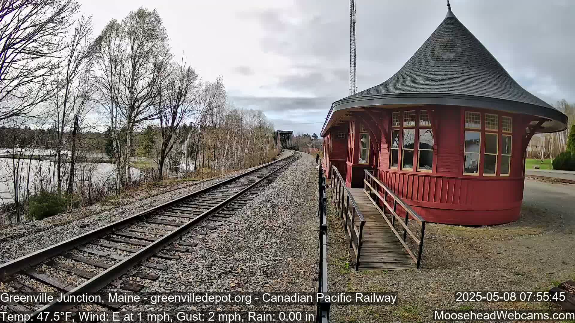 A red, round train depot with a conical roof sits beside train tracks that curve into the distance under a mostly cloudy sky, with a body of water and bare trees visible in the background.