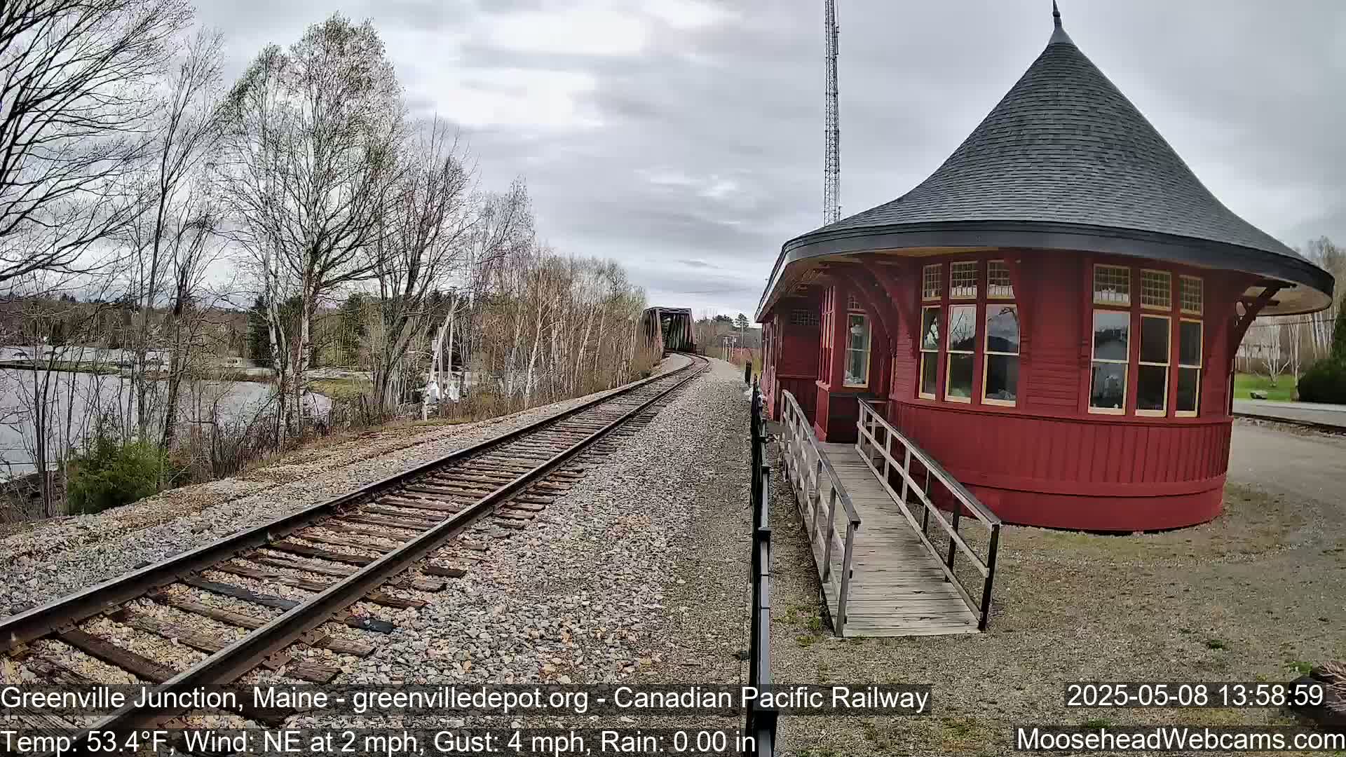 A red, round train depot with a ramp sits next to railroad tracks curving toward a bridge under an overcast sky.
