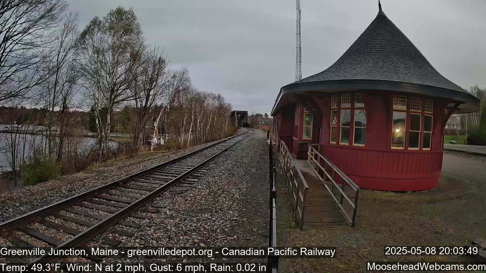 A red, round train station with a conical roof sits beside train tracks leading to a bridge, under a gray, rainy sky.