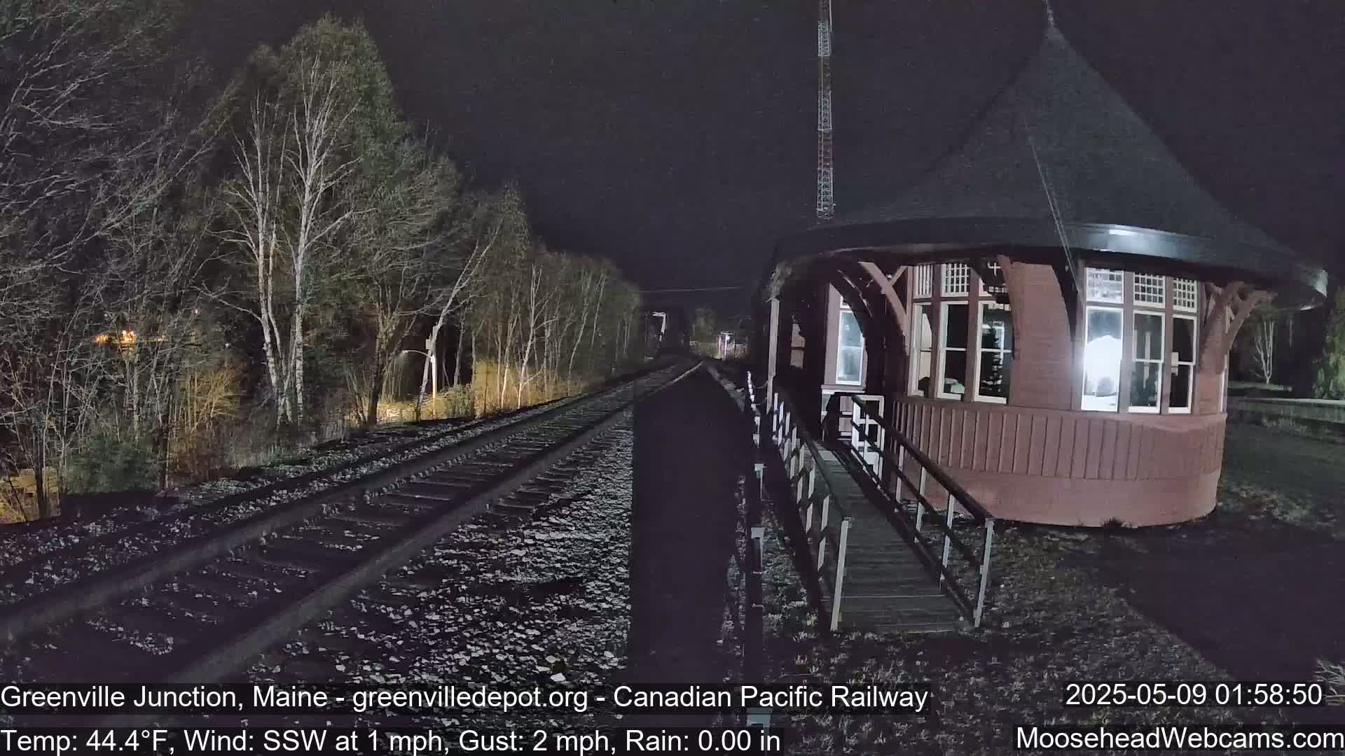 A red octagonal train signal house is situated beside railroad tracks at night during light rain, with bare trees lining the tracks in the background.
