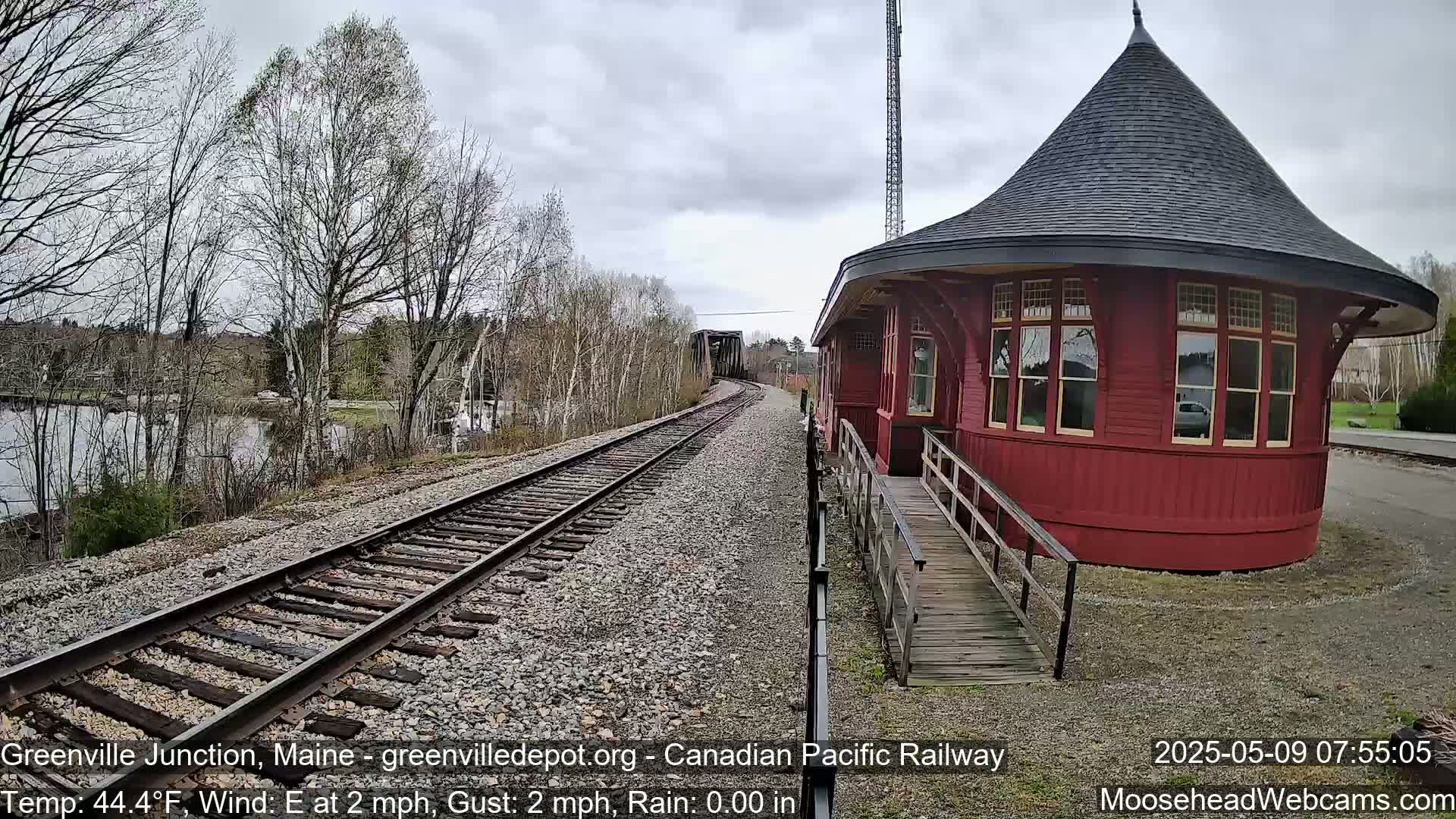 A red, round train depot with a conical roof sits beside train tracks curving towards a bridge, under a mostly cloudy sky.