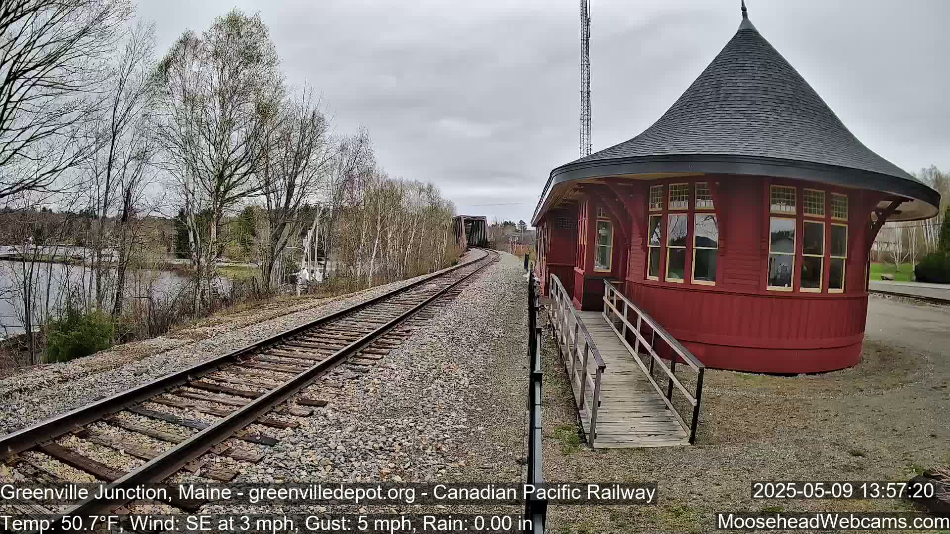 A red, round train depot with a conical roof sits beside railroad tracks curving toward a bridge, under an overcast sky.