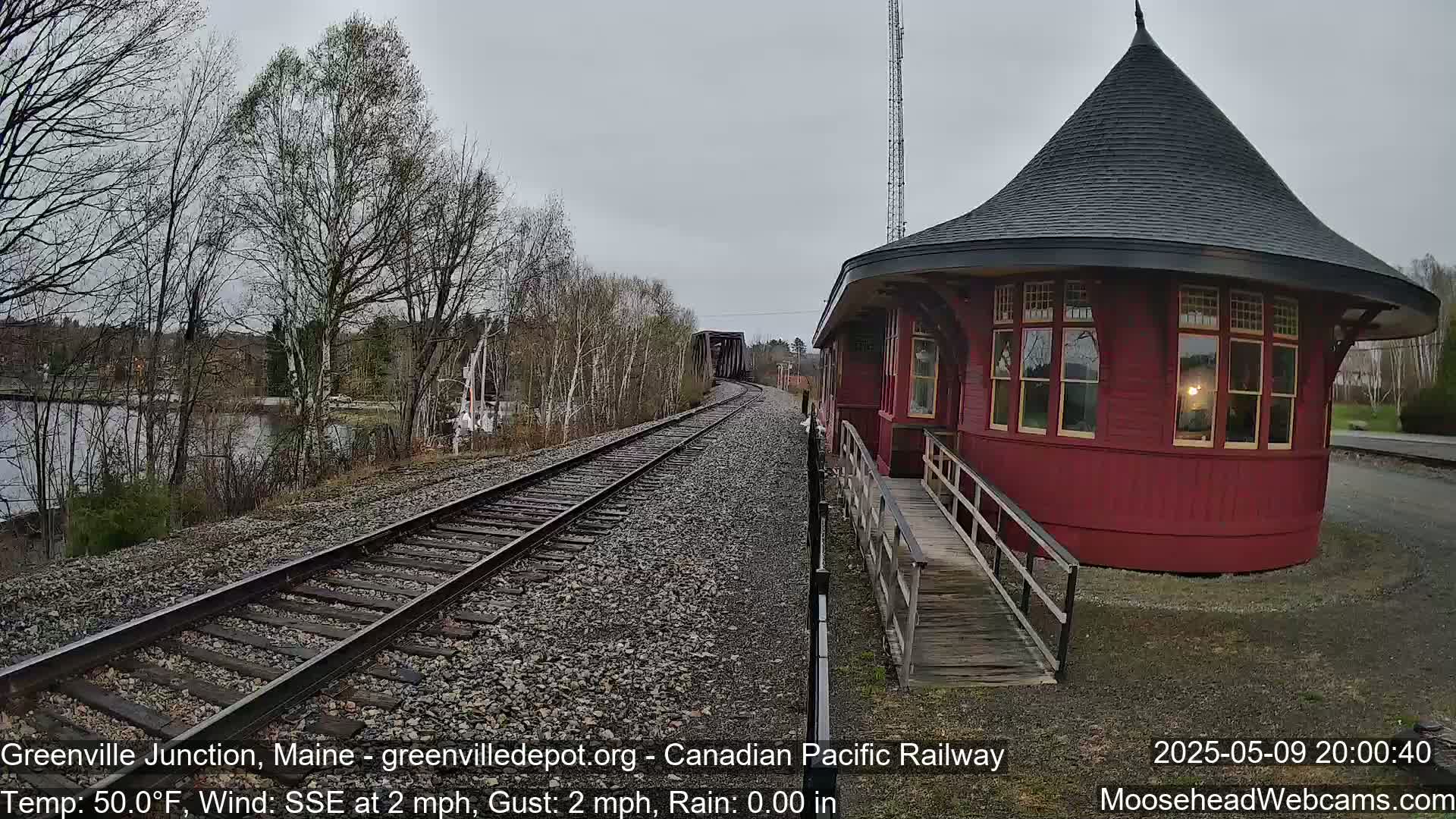 A red, round train station with a dark roof stands next to railway tracks that curve toward a distant bridge under an overcast sky.
